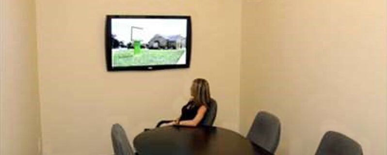 A woman sitting in a chair at the end of a conference room table looking at the TV that is on the wall in Hagerstown, Maryland offers court transcription services and more